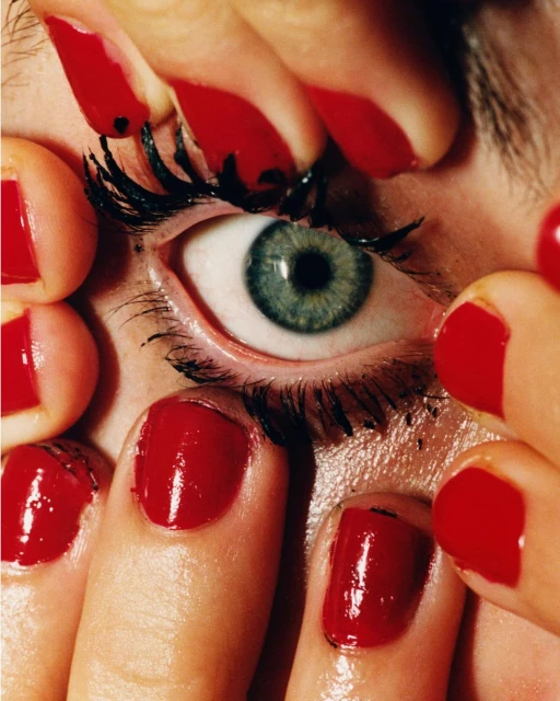 Macro shot of an eye surrounded by fingers with glossy red nails, capturing detail of beauty, skincare, and cosmetic elements.
