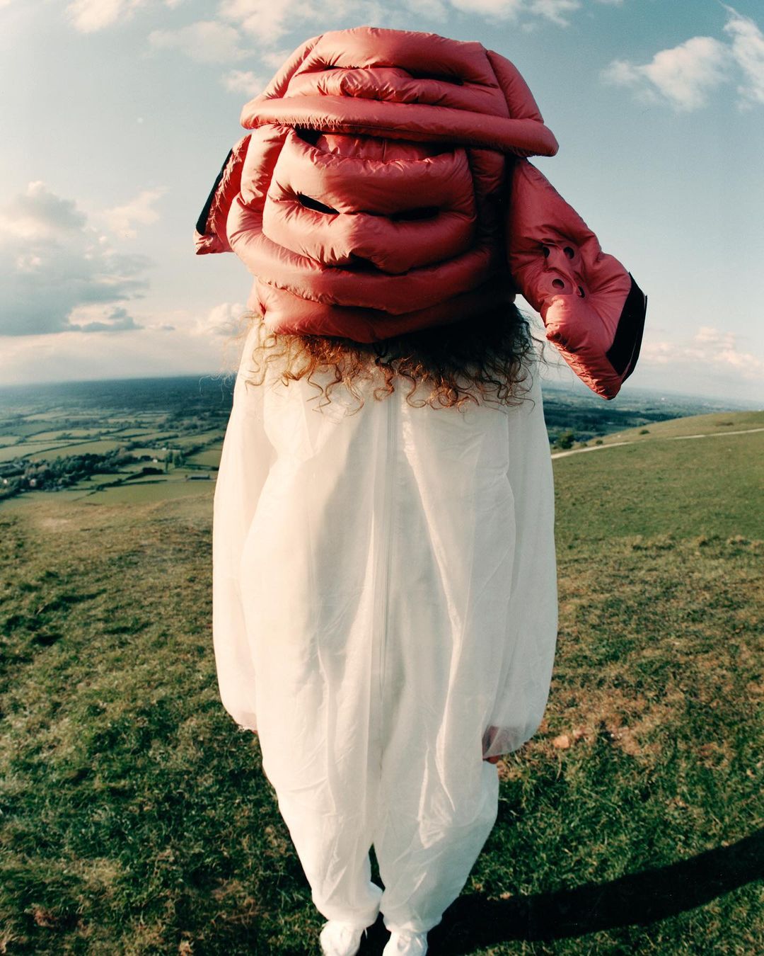 Person wearing a pink puffer jacket over their head, standing in a white jumpsuit in a grassy, hilly landscape under a cloudy sky.
