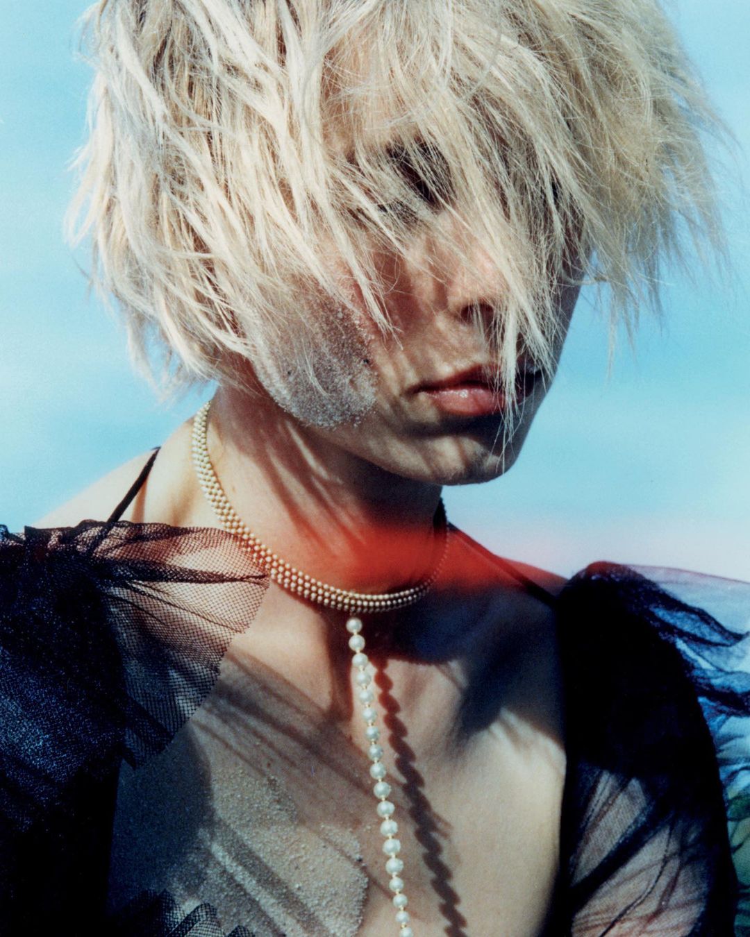 Close-up portrait of an adult female with blonde hair, pale skin, a pearl necklace, and a sheer black top against a blue background.
