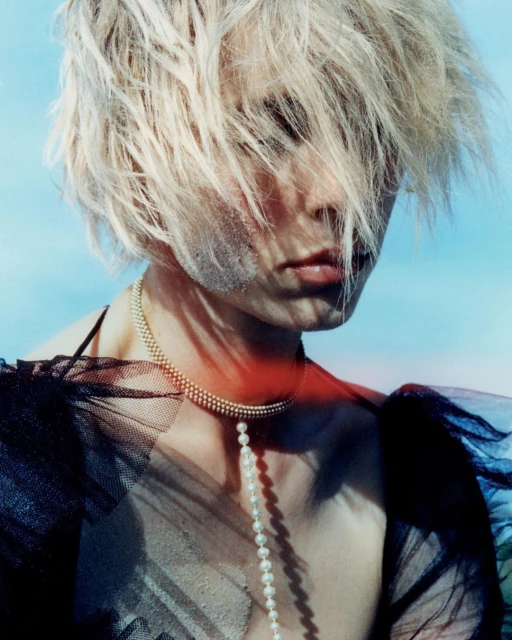 Close-up portrait of an adult female with blonde hair, pale skin, a pearl necklace, and a sheer black top against a blue background.