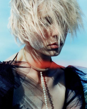 Close-up portrait of an adult female with blonde hair, pale skin, a pearl necklace, and a sheer black top against a blue background.