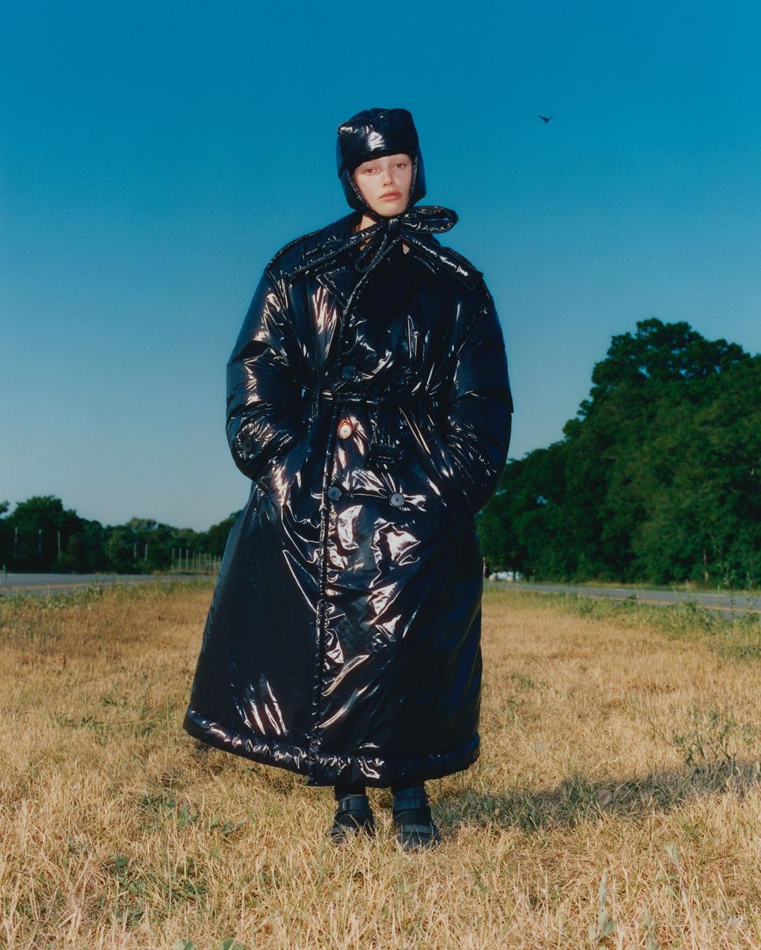 Model wears shiny black puffer coat and hat in an outdoor fashion shot. Grass and trees form the background under a clear blue sky.