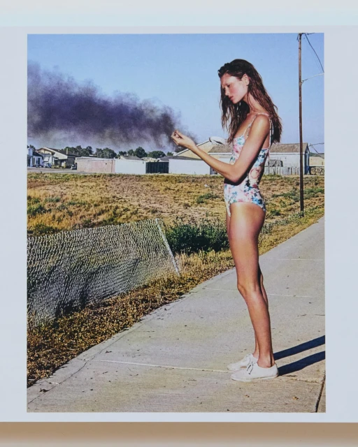 Woman in floral swimsuit stands on concrete path in sun with dark smoke plume rising from distant field against blue sky backdrop.