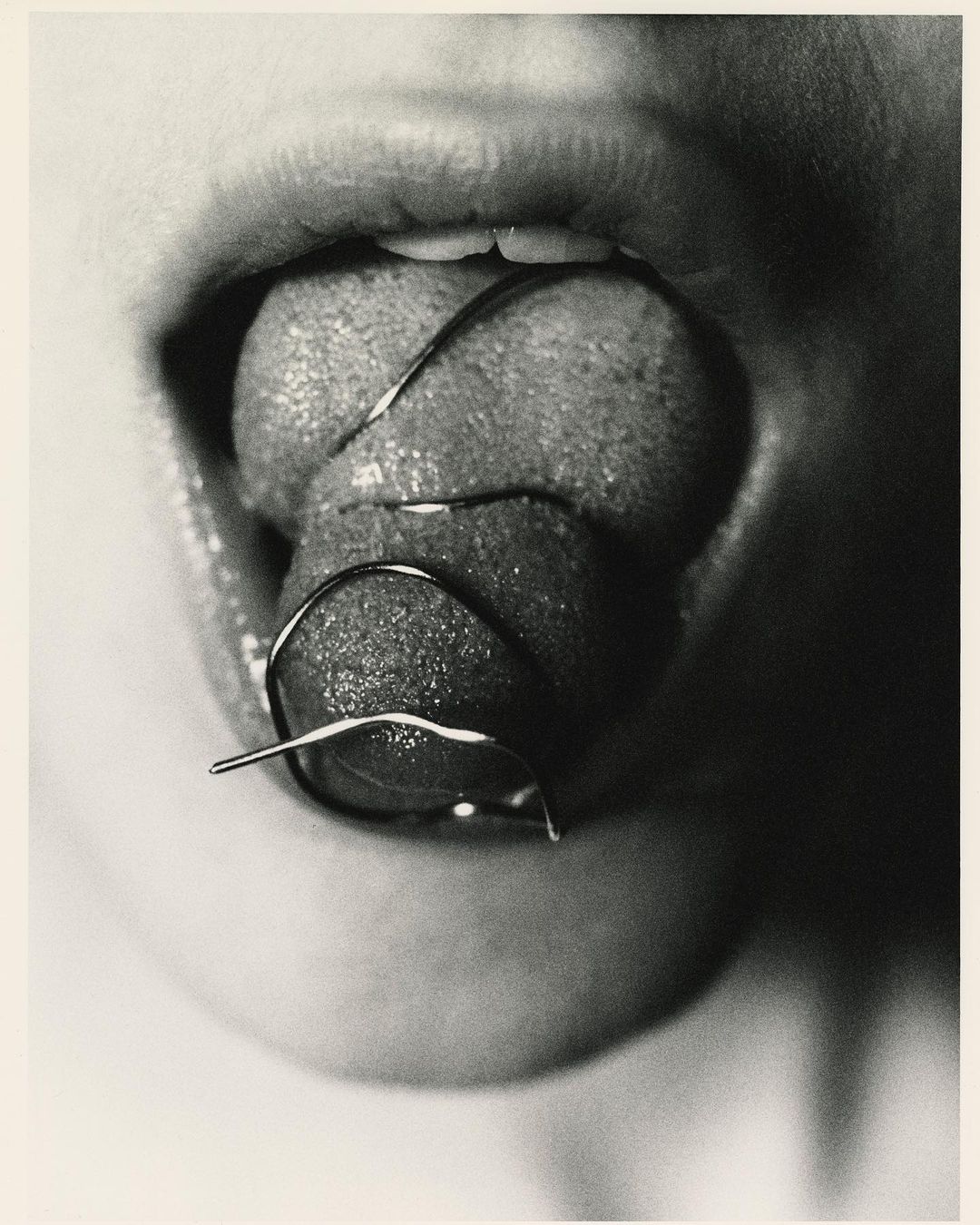 Monochrome close-up of a person with their mouth open and tongue sticking out, with a metal dental device on the tongue.
