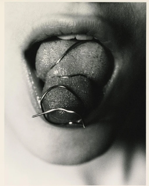 Monochrome close-up of a person with their mouth open and tongue sticking out, with a metal dental device on the tongue.