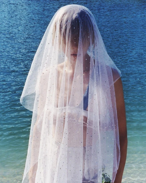 Woman in a sparkly white tulle veil stands in front of a blue ocean with fair skin, creating an ethereal and bright composition.