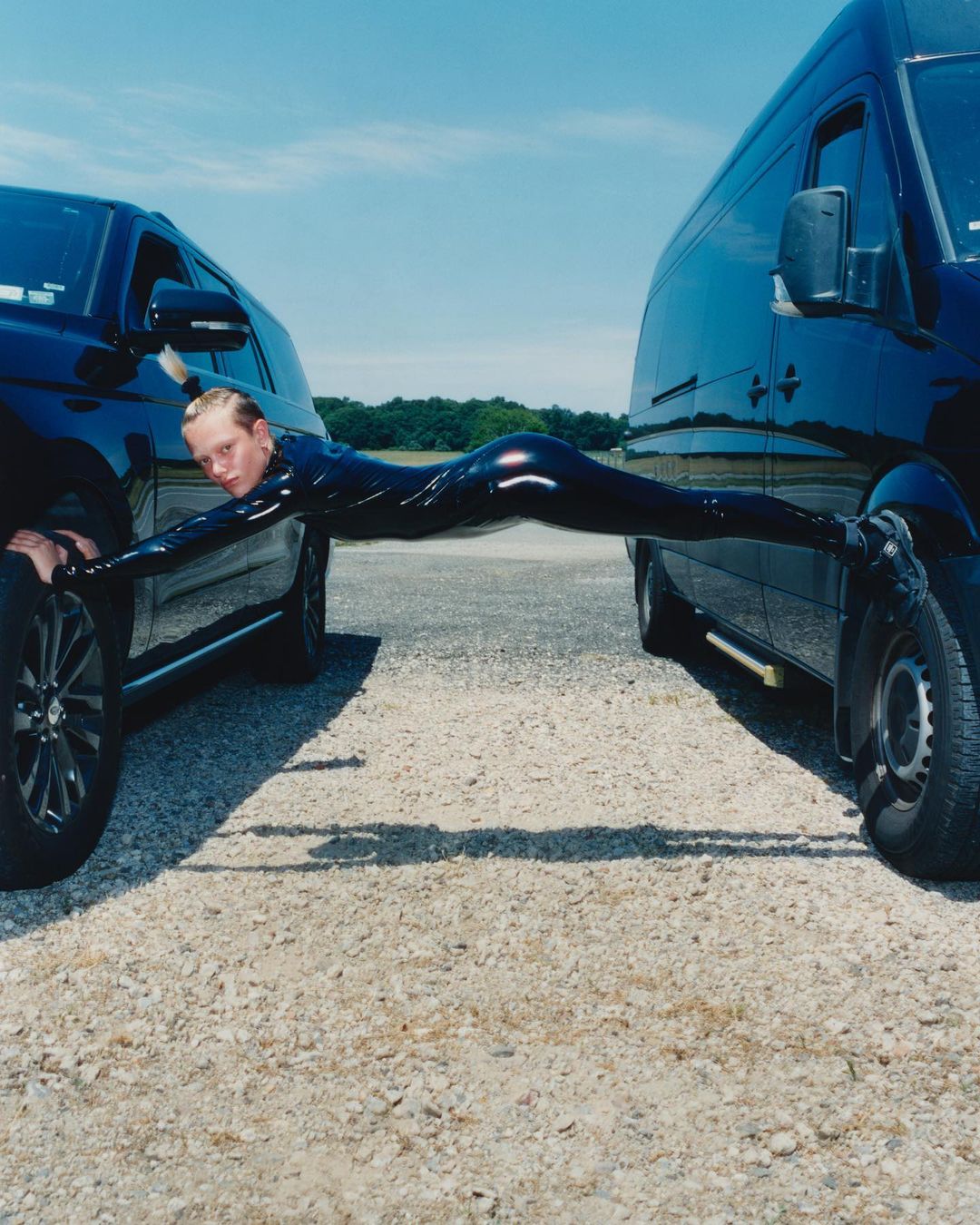Person in black catsuit performing a split between two black SUVs, set against a clear blue sky and a gravel road surface.