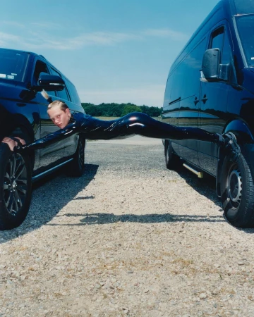 Person in black catsuit performing a split between two black SUVs, set against a clear blue sky and a gravel road surface.
