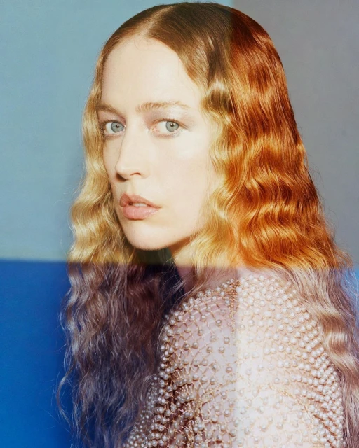 Portrait of a beautiful woman with long auburn wavy hair and wearing a pearled garment, posing against a blue and gray background.