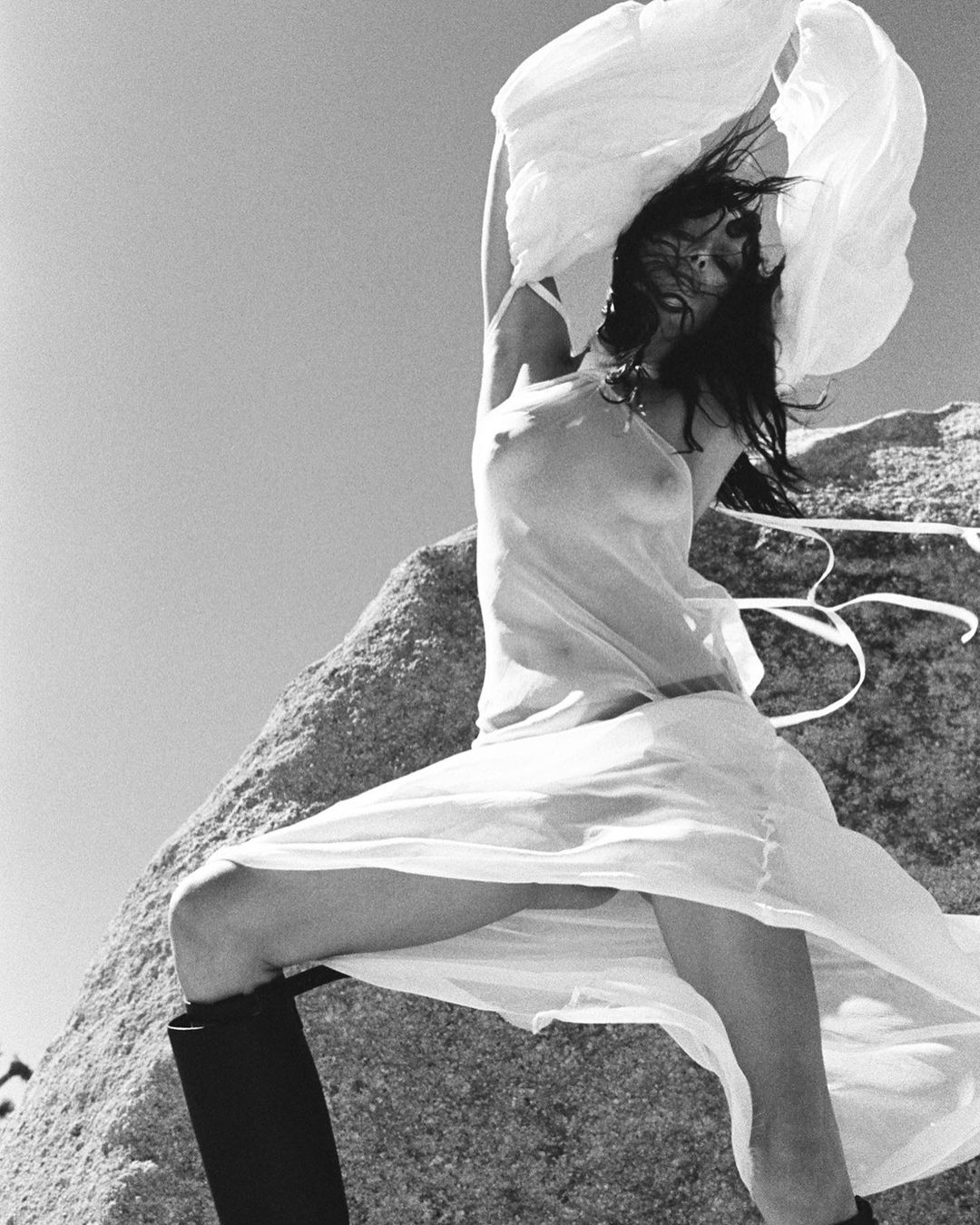 Model in sheer white dress posing on a rocky outcrop outdoors photographed by Drew Vickers in black and white.