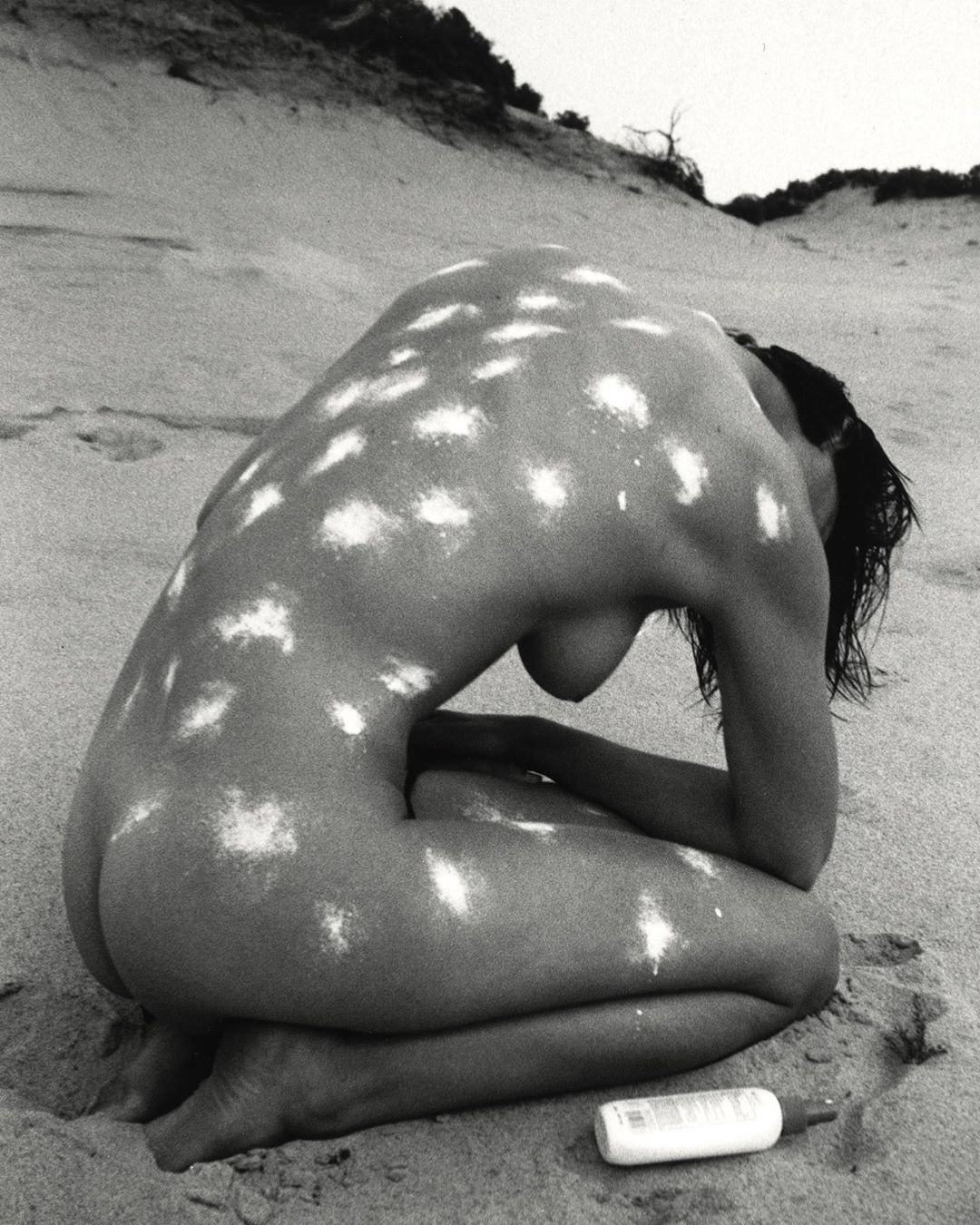Monochrome nude photograph of an adult woman kneeling on a beach with white dots covering her skin, with sand dunes behind her.