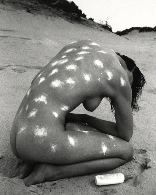 Monochrome nude photograph of an adult woman kneeling on a beach with white dots covering her skin, with sand dunes behind her.