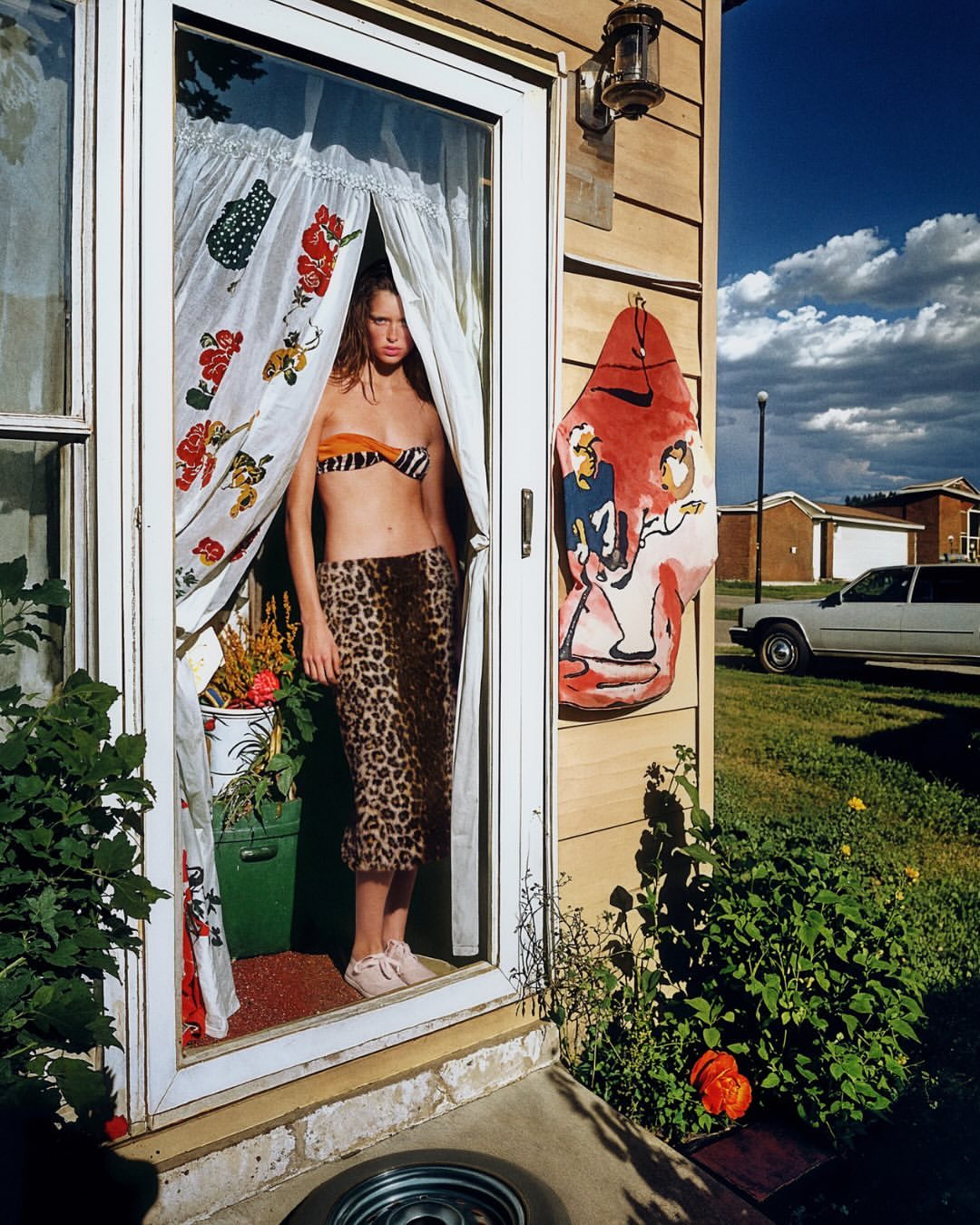 A woman stands in a doorway wearing a bikini top and leopard print skirt, with a white door, floral curtain, and suburb in the background.