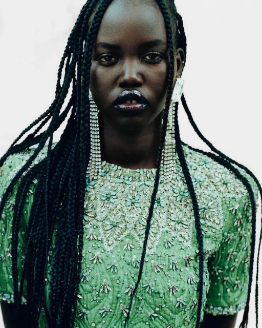 Portrait of a dark-skinned woman with braids, wearing a green beaded dress and long crystal earrings, against a pale background.