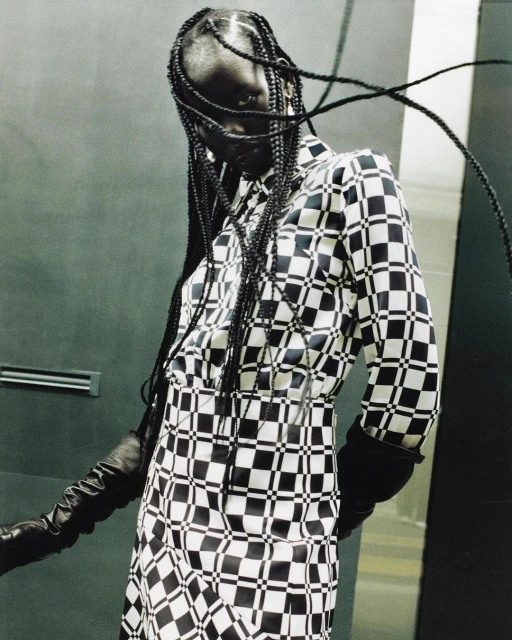 A dark-skinned person poses in a black-and-white checkerboard dress and gloves, set against a muted green background.