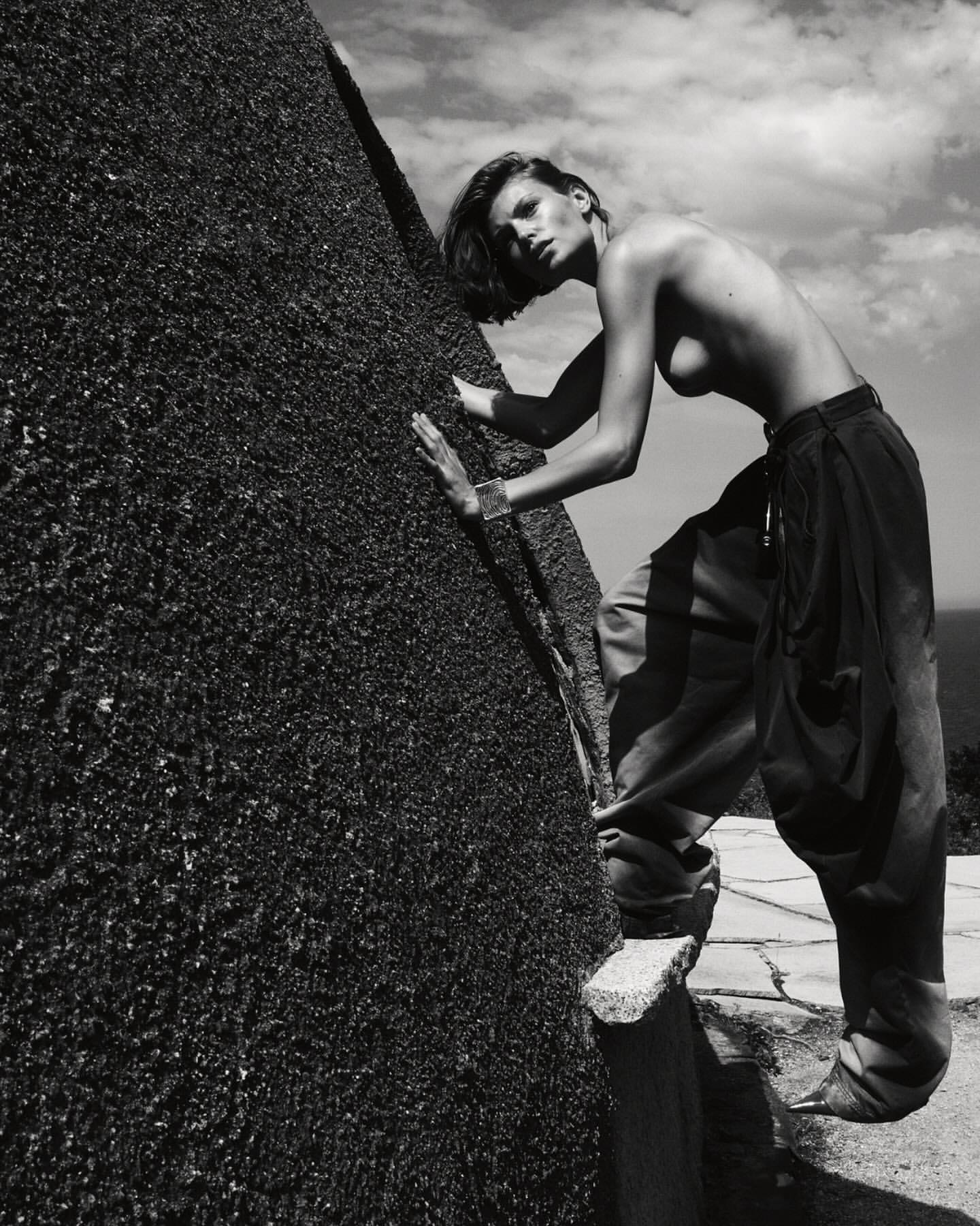 Monochrome image of a woman in loose trousers posing next to a rough stone structure under a cloudy sky, appearing to be outdoors.