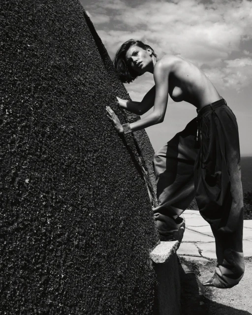 Monochrome image of a woman in loose trousers posing next to a rough stone structure under a cloudy sky, appearing to be outdoors.