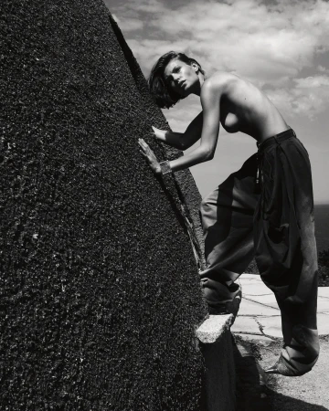 Monochrome image of a woman in loose trousers posing next to a rough stone structure under a cloudy sky, appearing to be outdoors.
