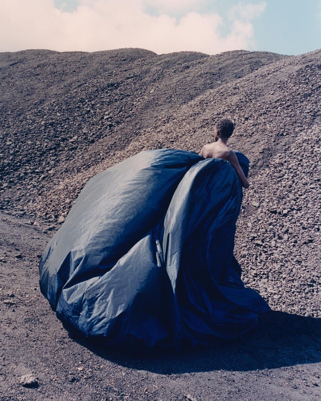 Back view of an adult male wearing a voluminous dark draped fabric, posed against a background of earth and rocky terrain under a cloudy sky.