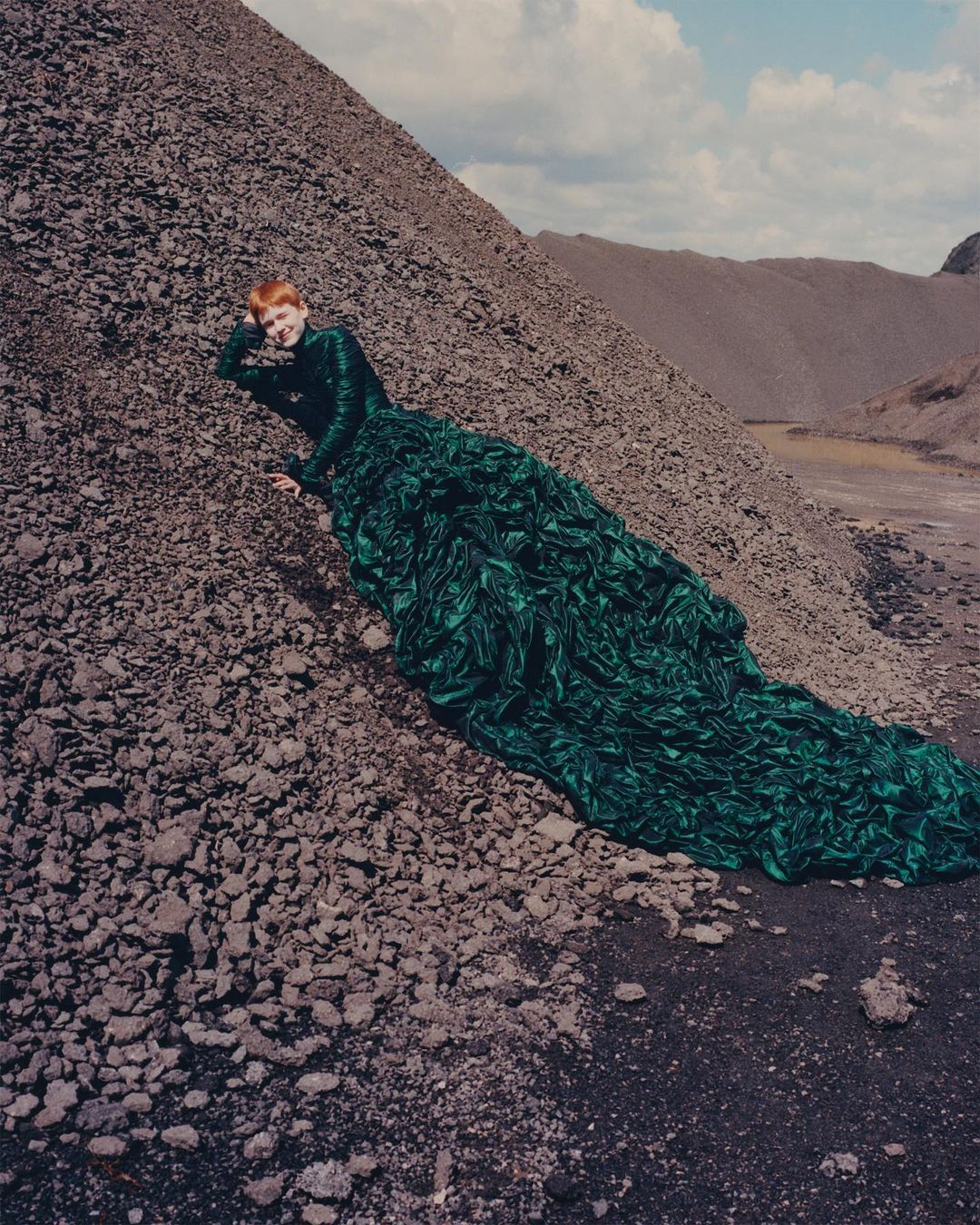 A woman in a green gown reclines on a gray rocky hill under a cloudy sky, showcasing a blend of high fashion and stark, natural landscape.