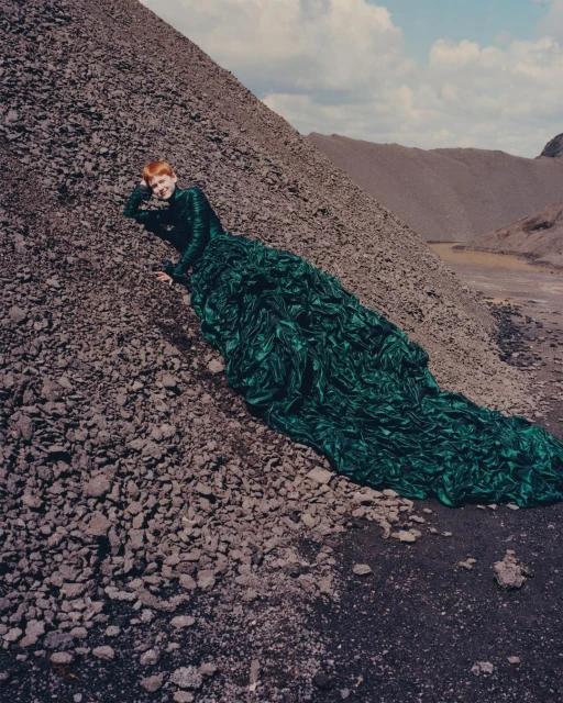 A woman in a green gown reclines on a gray rocky hill under a cloudy sky, showcasing a blend of high fashion and stark, natural landscape.