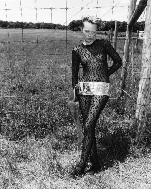 A woman in a leopard print bodysuit poses near a wire fence in a rural setting. She is wearing a wide, reflective belt accessory.