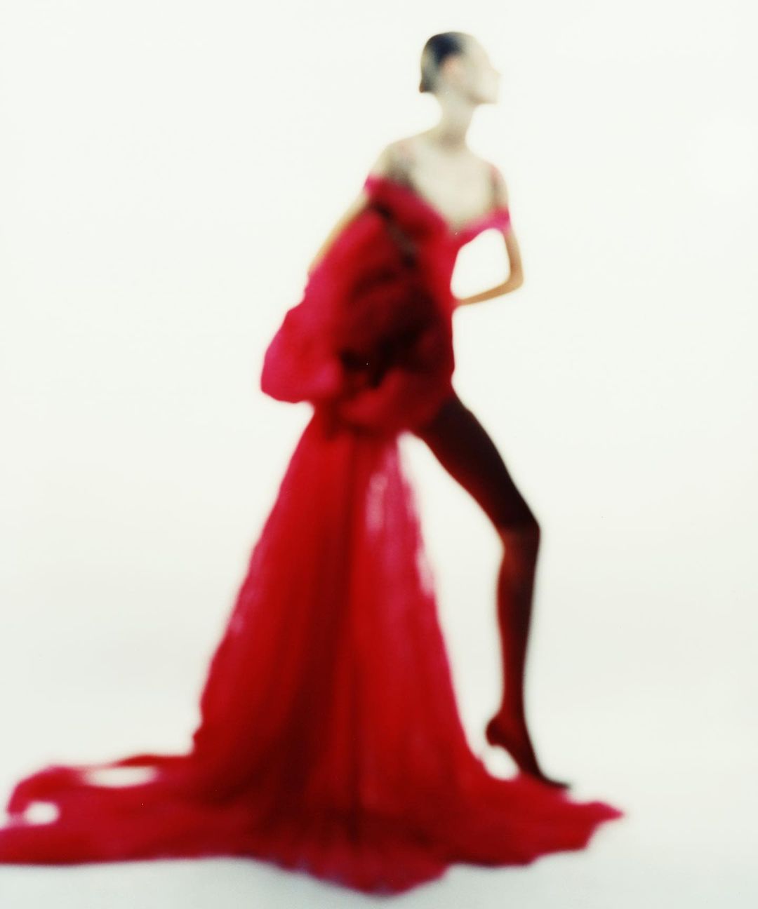 Fashion model posing in a dramatic red gown with an off-the-shoulder neckline against a bright white backdrop in a studio setting.
