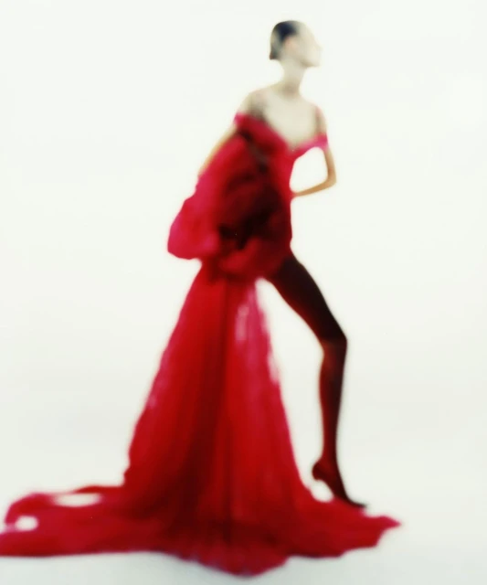 Fashion model posing in a dramatic red gown with an off-the-shoulder neckline against a bright white backdrop in a studio setting.