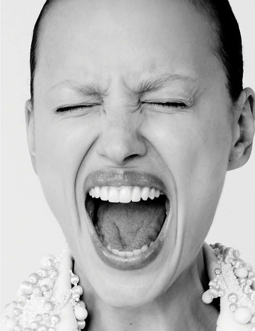 Close-up black and white portrait of a young woman screaming with her eyes closed, wearing a beaded collar and standing against a white background.