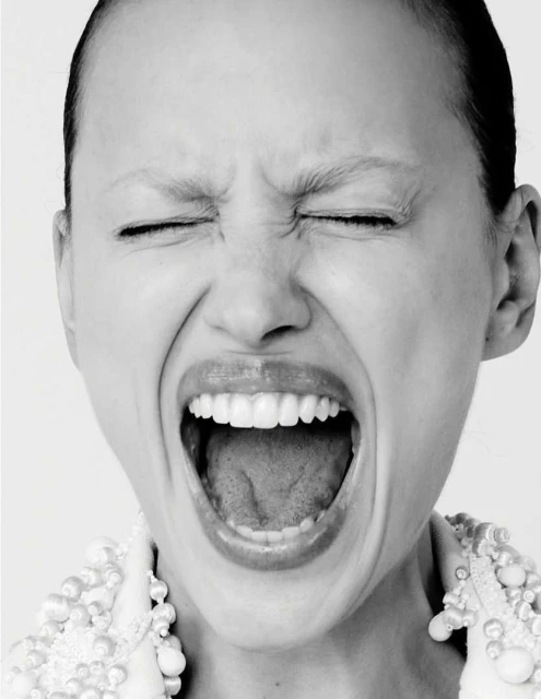 Close-up black and white portrait of a young woman screaming with her eyes closed, wearing a beaded collar and standing against a white background.