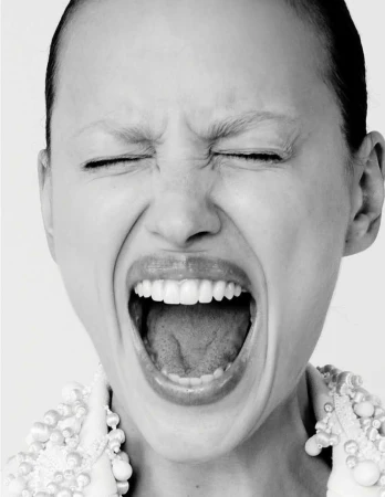 Close-up black and white portrait of a young woman screaming with her eyes closed, wearing a beaded collar and standing against a white background.