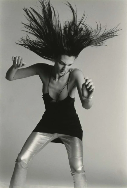 Black and white portrait of a woman with long hair tossed in the air, wearing a black tank top and shiny silver pants in a studio.