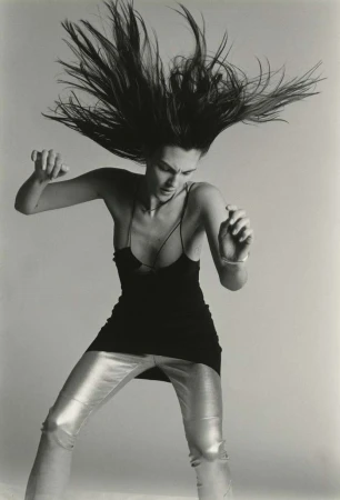 Black and white portrait of a woman with long hair tossed in the air, wearing a black tank top and shiny silver pants in a studio.