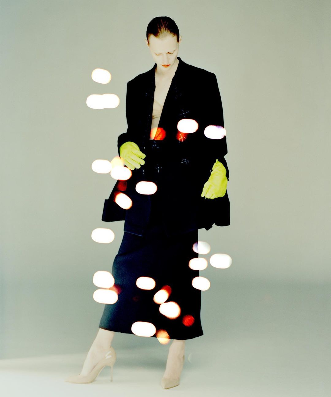 A tall fashion model with pale skin poses in a black suit, neon gloves, and beige high heels against a soft gray background.