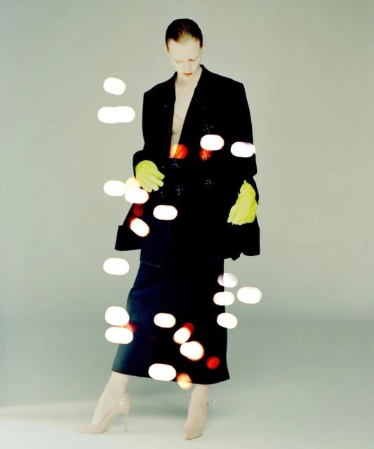 A tall fashion model with pale skin poses in a black suit, neon gloves, and beige high heels against a soft gray background.