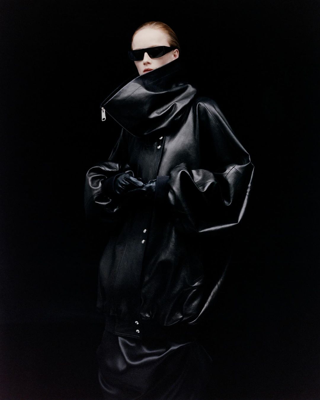 Model wearing dark sunglasses and an oversized black leather jacket with a high collar against a dark background