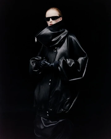 Model wearing dark sunglasses and an oversized black leather jacket with a high collar against a dark background