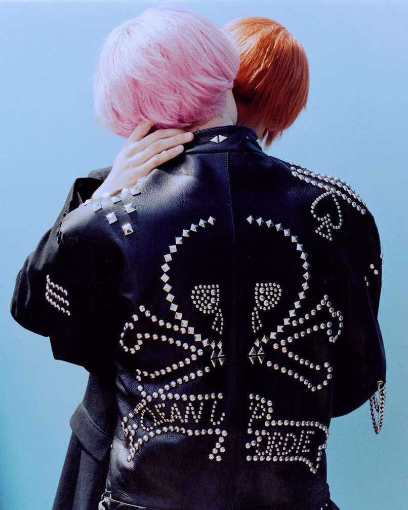 Two figures embrace, backs to camera, punk-style leather jacket adorned with skull and crossbones and 'Clean Up or Die' slogan against a light blue backdrop.