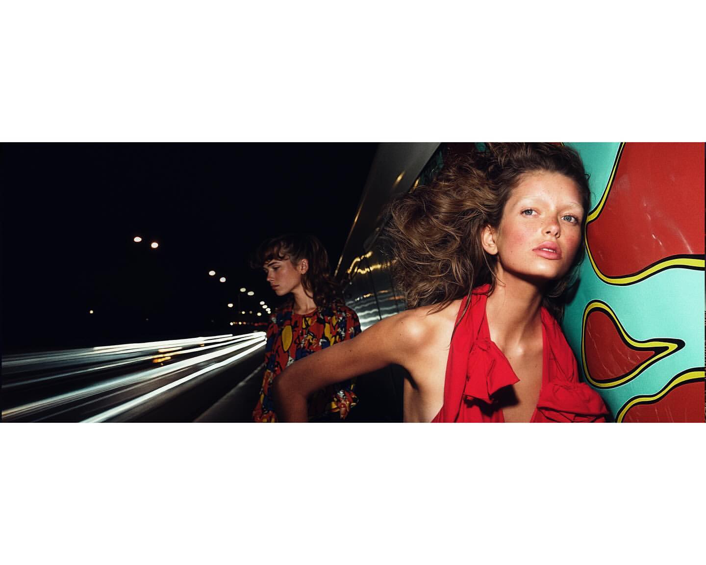 Two fashion models pose at night on a highway overpass. One wears a red dress and the other wears a colorful patterned top.