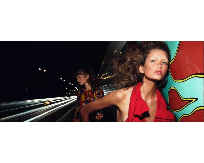 Two fashion models pose at night on a highway overpass. One wears a red dress and the other wears a colorful patterned top.