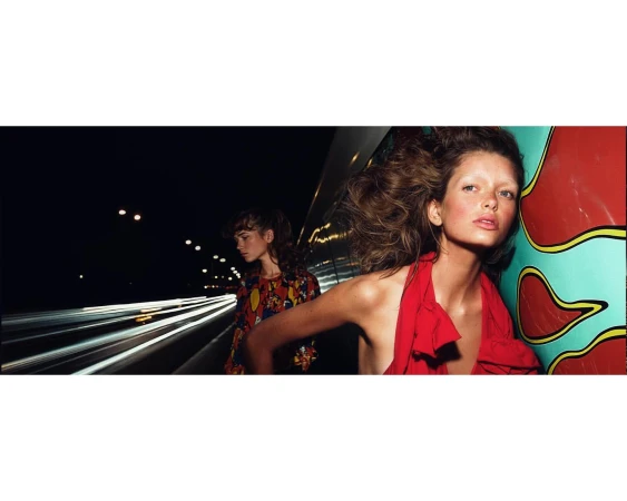 Two fashion models pose at night on a highway overpass. One wears a red dress and the other wears a colorful patterned top.
