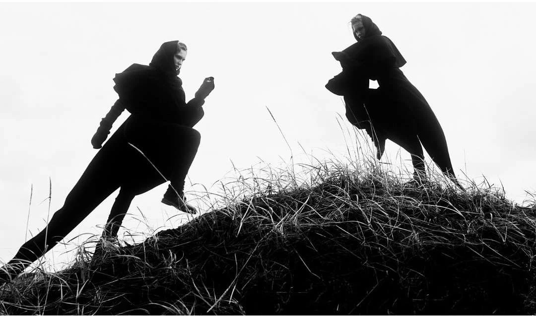 Two figures in dark cloaks traverse a grassy hilltop against a bright, overcast sky in a black-and-white, low-angle outdoor shot.