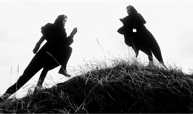 Two figures in dark cloaks traverse a grassy hilltop against a bright, overcast sky in a black-and-white, low-angle outdoor shot.