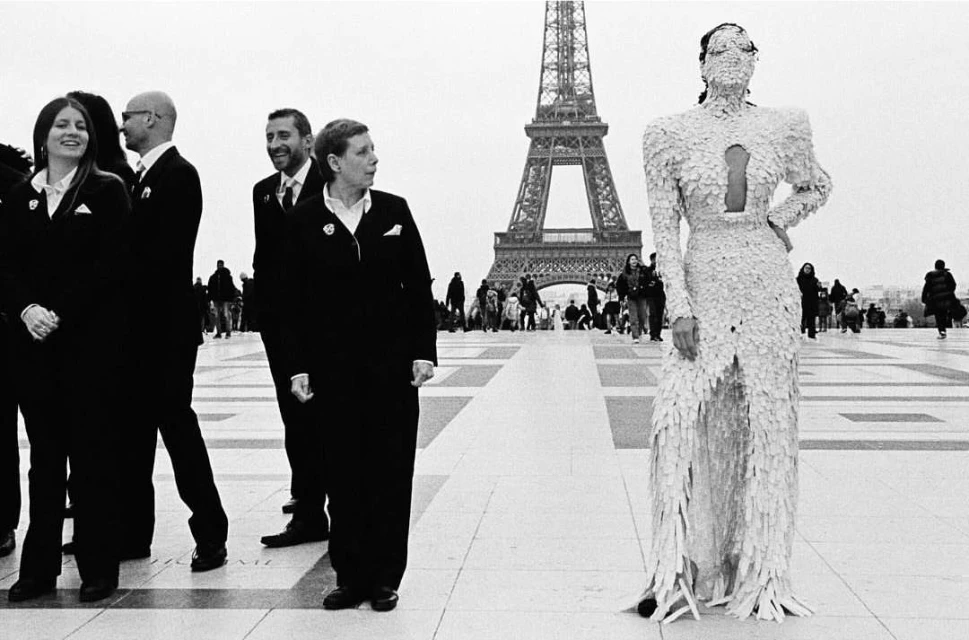 Fashion show in Paris with a model in a textured dress and a group in black suits with the Eiffel Tower in the background.