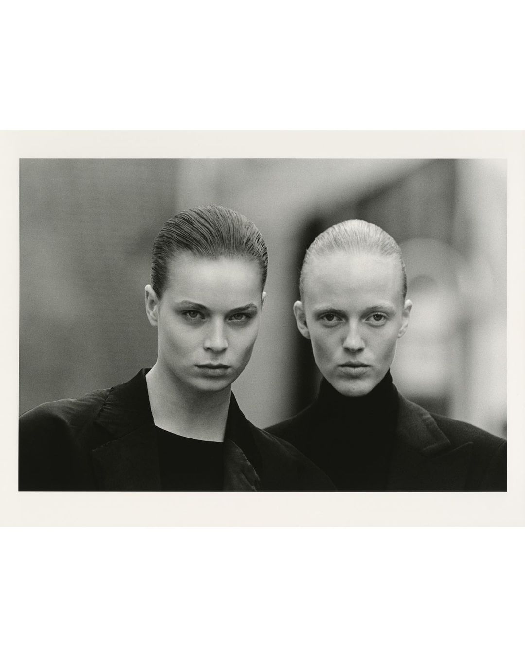 Monochrome portrait of two women in black jackets, close-up shot with blurred background, serious expressions, and slicked-back hairstyles.