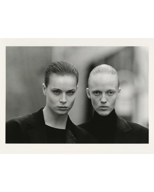 Monochrome portrait of two women in black jackets, close-up shot with blurred background, serious expressions, and slicked-back hairstyles.