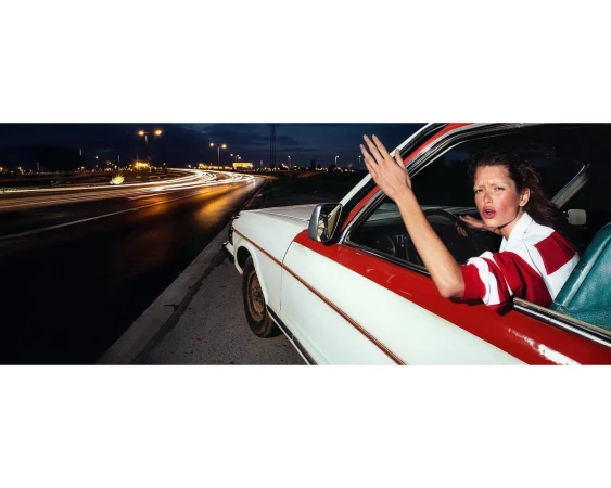Woman in a white car with a red stripe waves her hand at blurred street lights at night, showing emotion and retro style.