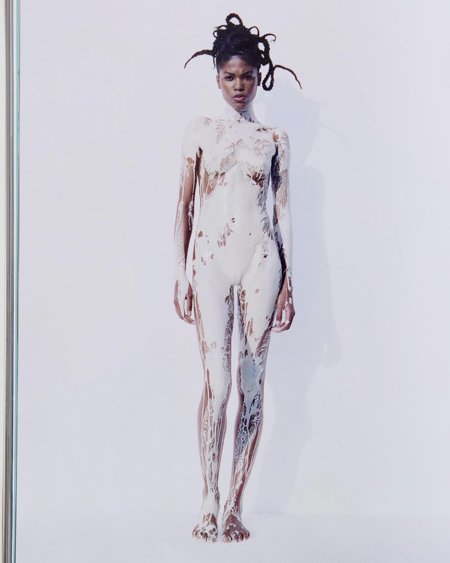 Full-length studio shot of an adult woman covered in white paint, standing against a white background. The woman has dark skin and dreadlocks.