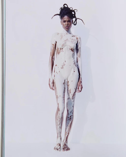 Full-length studio shot of an adult woman covered in white paint, standing against a white background. The woman has dark skin and dreadlocks.
