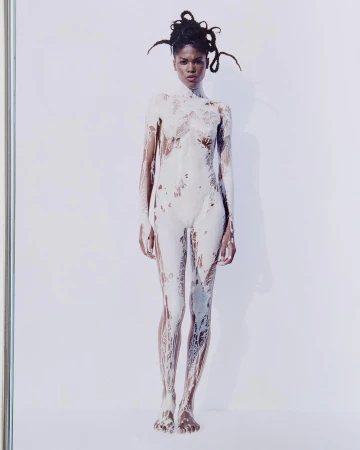 Full-length studio shot of an adult woman covered in white paint, standing against a white background. The woman has dark skin and dreadlocks.